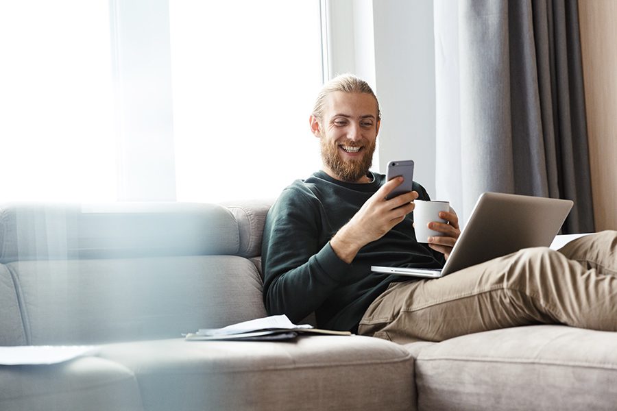Client Center - Young Man With Beard On Couch With Phone Cup of Coffee and Laptop